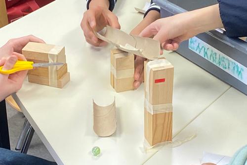 Mehrere Schülerhände bauen mit Holzklötzen, Papierrollen und Klebeband eine Murmelbahn. Auf dem Tisch liegt eine grüne Murmel, eine Schere ist im Einsatz.