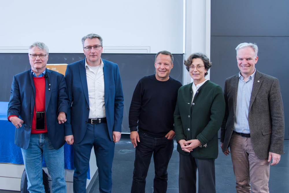 Fünf Männer und eine Frau posieren gemeinsam für ein Gruppenfoto in einem modernen Raum. Die Personen wirken freundlich und professionell, einige tragen Anzug, andere sind leger gekleidet. Im Hintergrund ist ein Tisch mit blauem Tuch zu sehen.