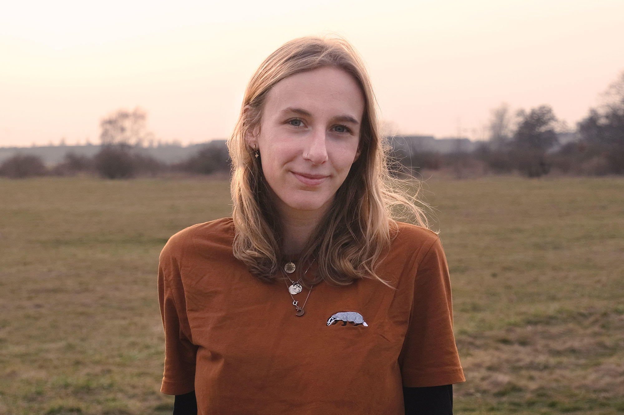 Britt Köhler, eine junge Frau mit schulterlangen blonden Haaren steht leicht lächelnd auf einer offenen Wiese bei sanftem Abendlicht. Sie trägt ein rostbraunes T-Shirt mit einem Dachs-Aufnäher und mehreren Ketten. Im Hintergrund sind Büsche und Bäume zu s