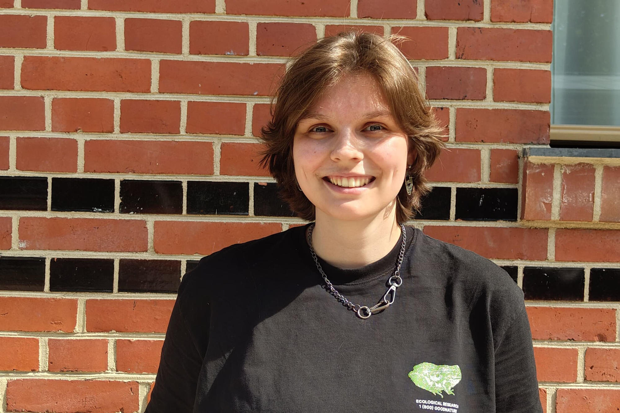 Jule Detmers, eine junge Frau mit kurzen, braunen Haaren, steht lächelnd vor einer Backsteinwand mit schwarzen Ziersteinen. Sie trägt ein schwarzes T-Shirt mit grünem Aufdruck, eine Halskette und hat die Hände in den Hosentaschen. Die Sonne scheint hell.