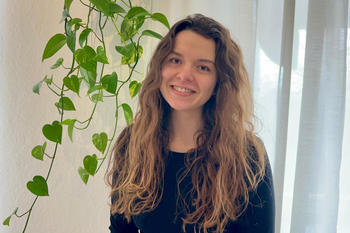 Theresa Langer, eine junge Frau mit langen, welligen braunen Haaren steht lächelnd vor einem hellen Vorhang. Neben ihr hängt eine grüne Rankpflanze in einem weißen Topf an der Wand. Das Licht wirkt weich und natürlich.